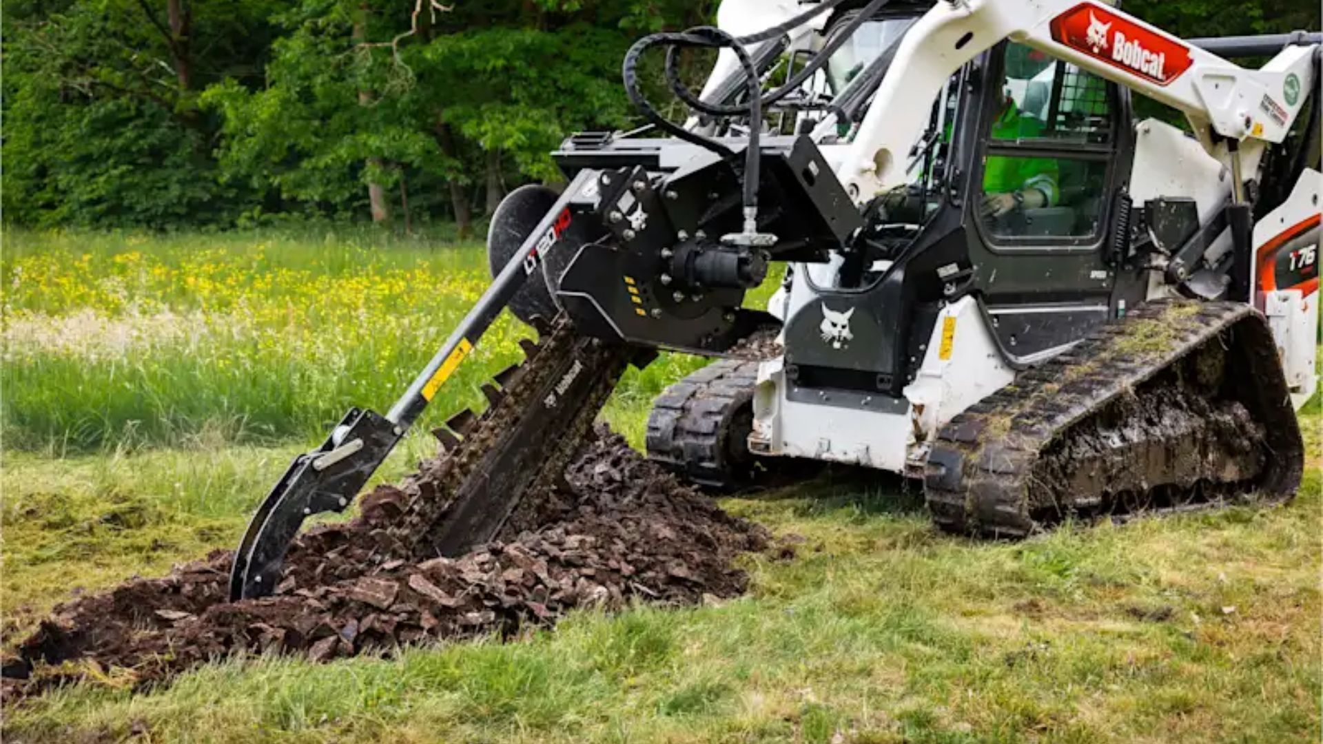 A tracked excavator with a digging attachment clearing muddy soil in a grassy field