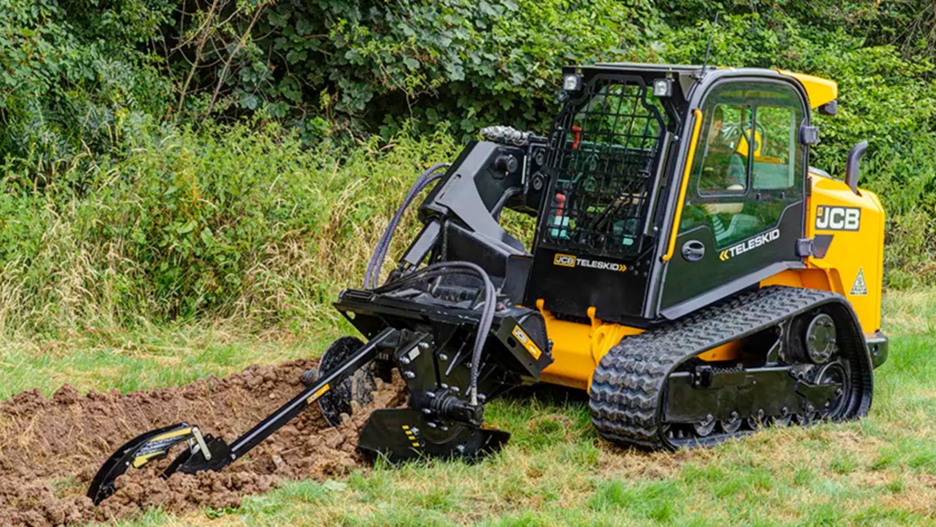 Yellow-and-black tracked JCB excavator mowing brush beside a grassy field and trees