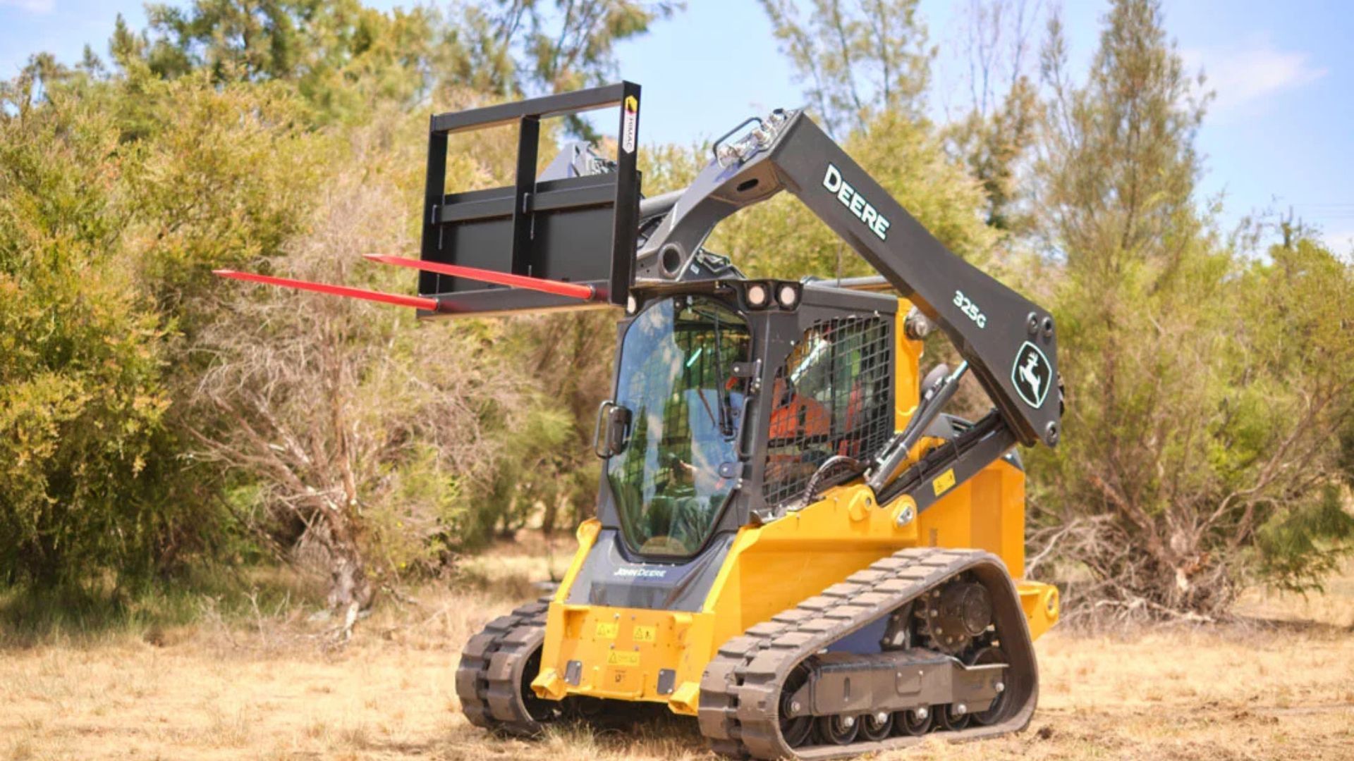 Yellow compact loader with raised forks on a dirt path near trees, outdoors