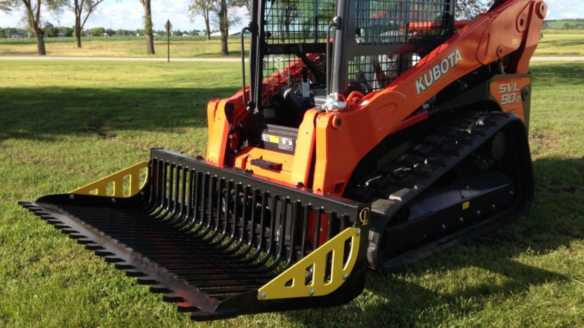 Orange skid-steer loader with front brush rake attachment on grass in a park