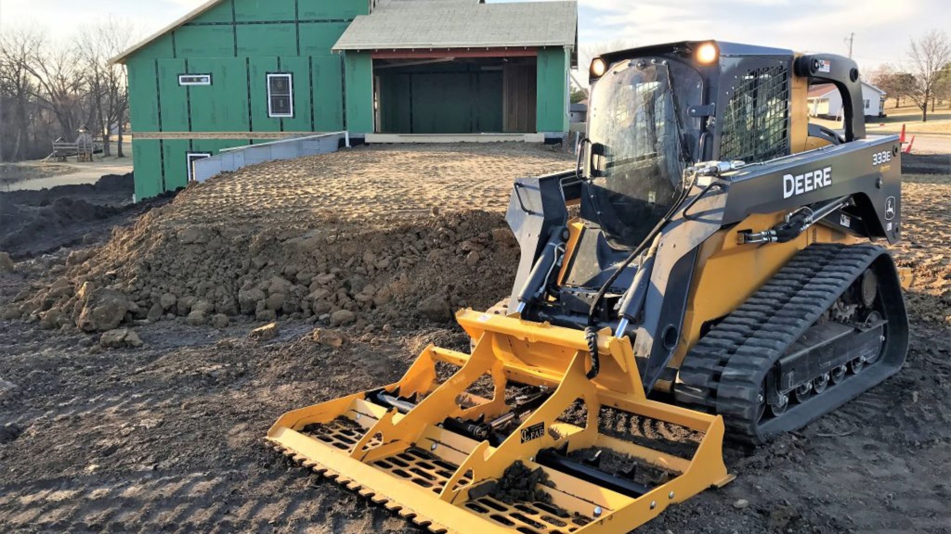 Yellow skid steer with grapple bucket on muddy ground beside a green building and dirt mound