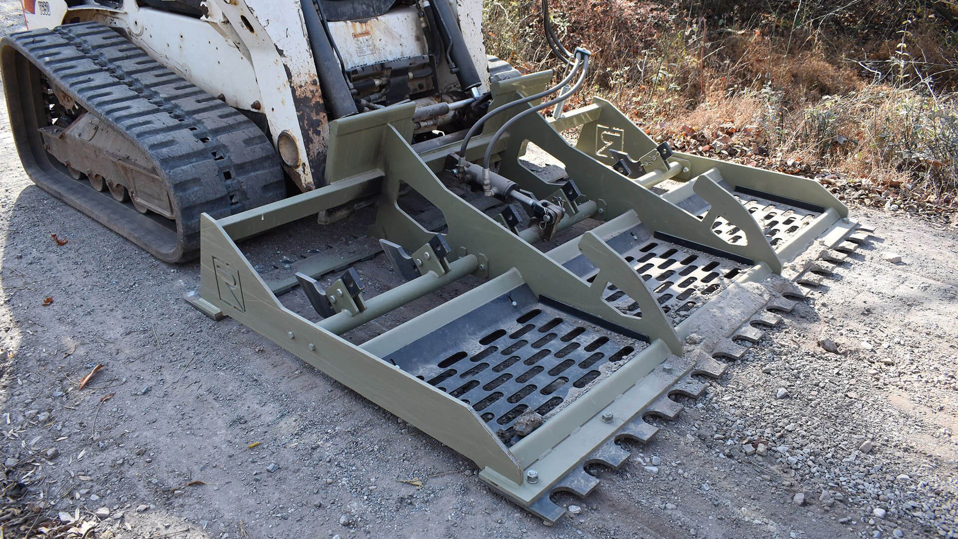 Tracked skid-steer attachment with metal grapple and raking tines on gravel ground