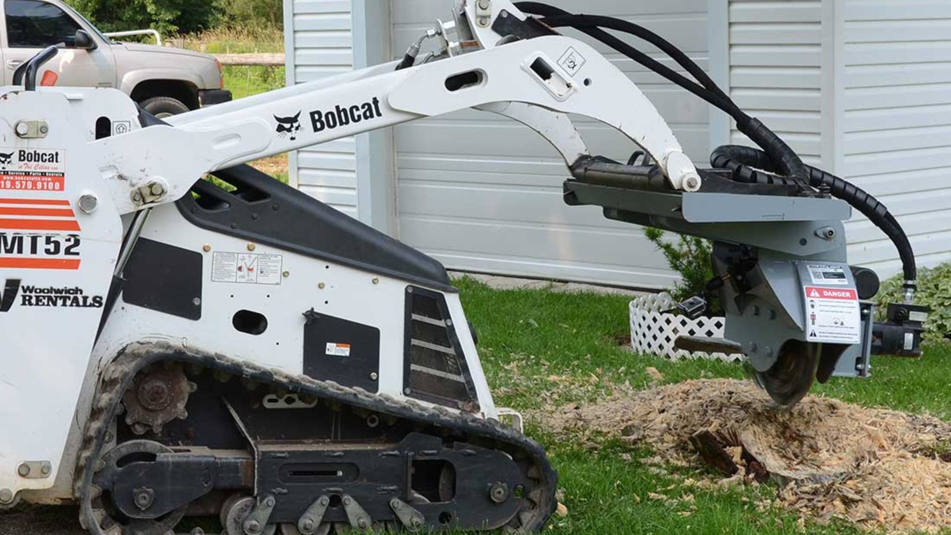 White Bobcat skid-steer loader scooping dirt beside a house with a front-mounted auger attachment