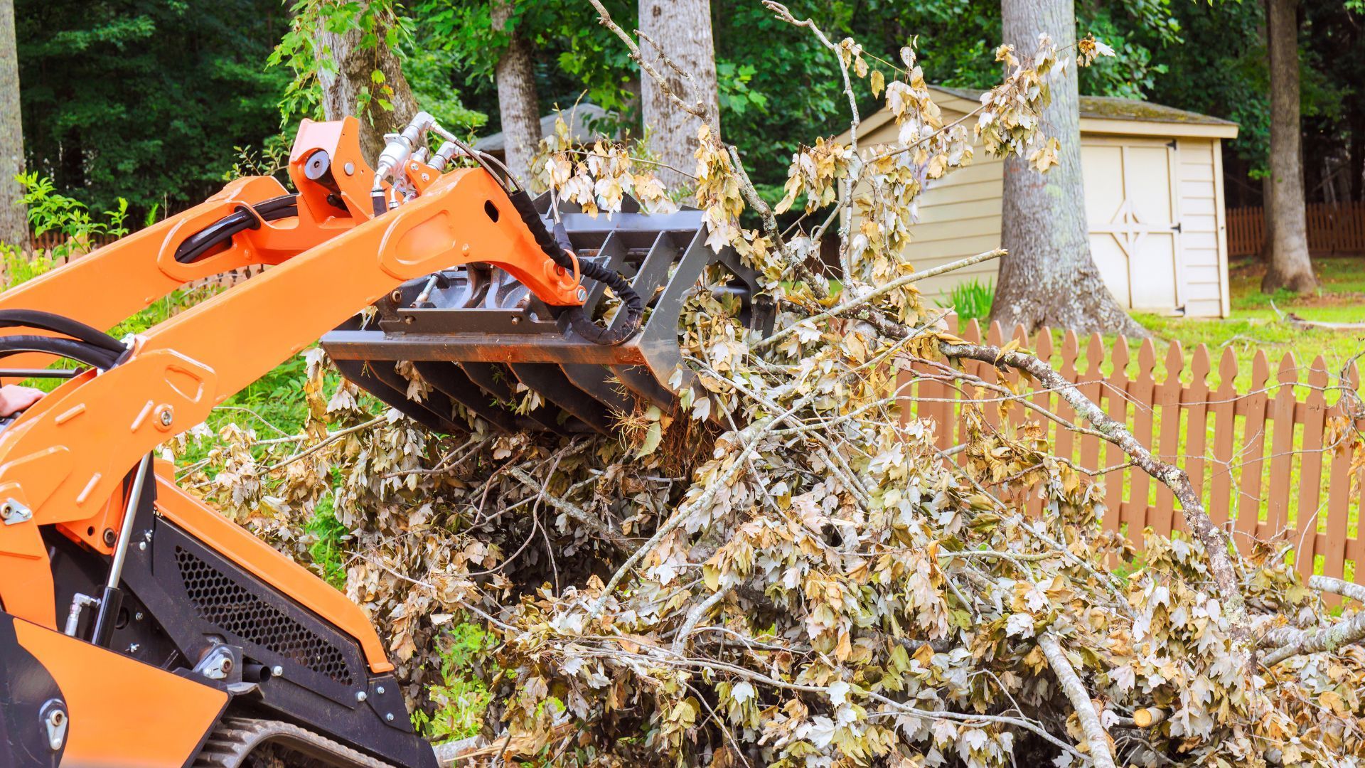 Orange excavator clearing a large pile of yard debris beside a fence and shed
