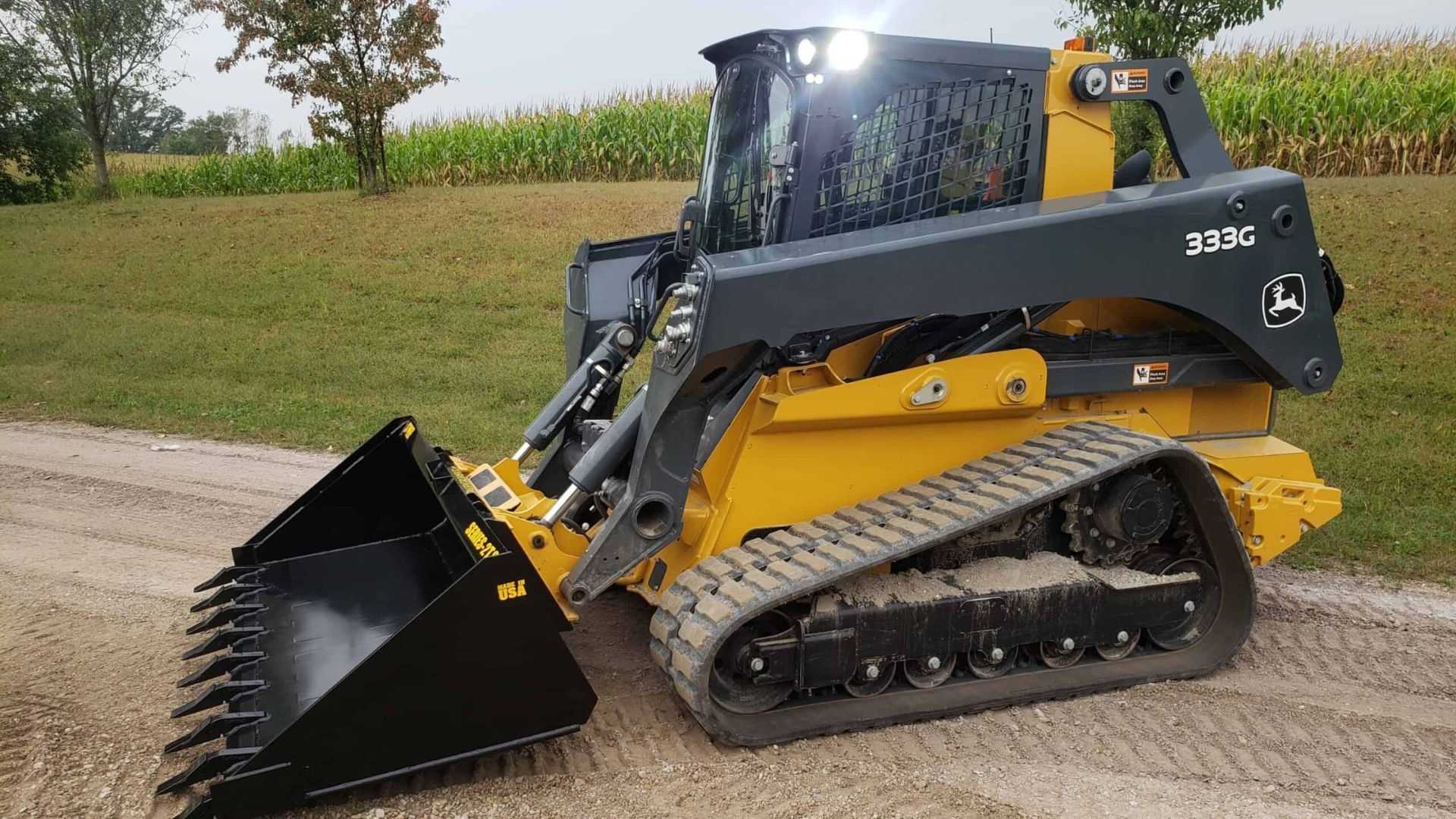 Yellow and black compact track loader with raised bucket on a gravel road beside a grassy field