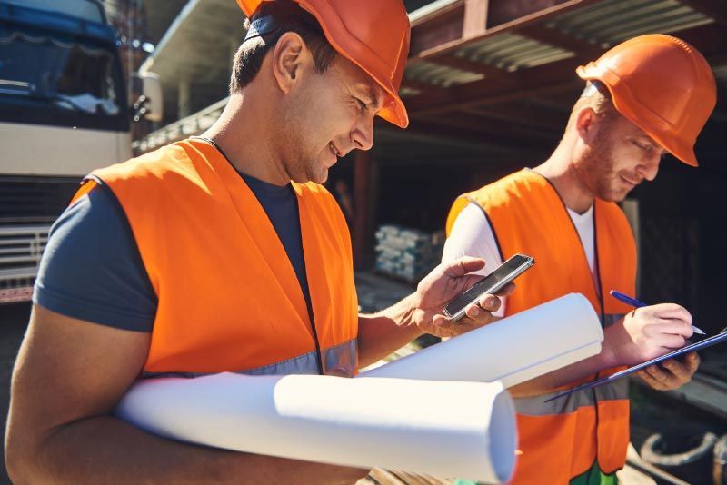 two construction workers are looking at a blueprint and one is using a cell phone .