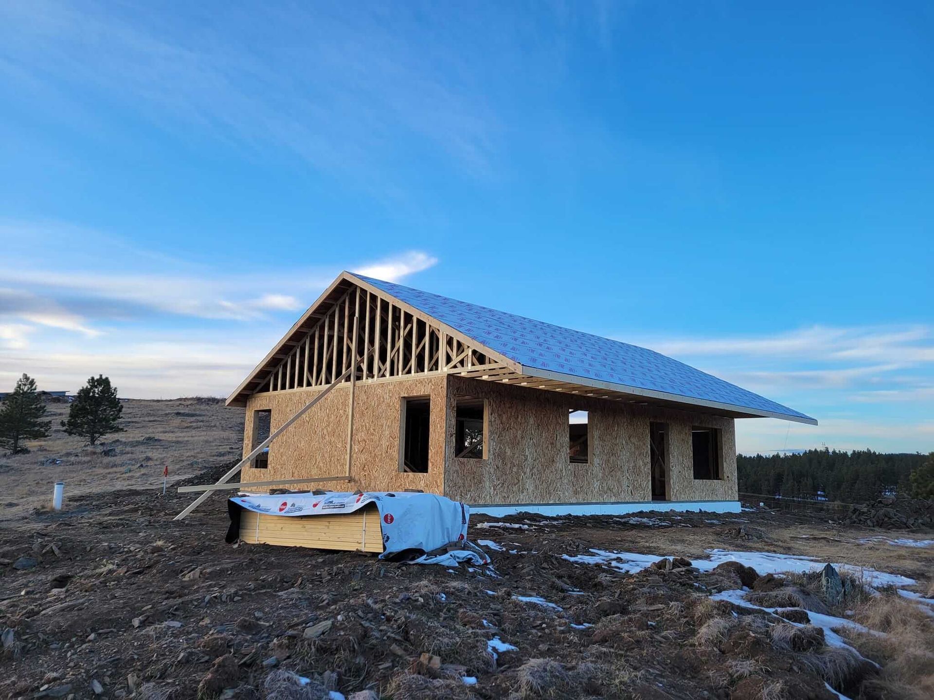 House under construction on a dirt lot; unfinished frame, plywood walls, metal roof, blue sky.