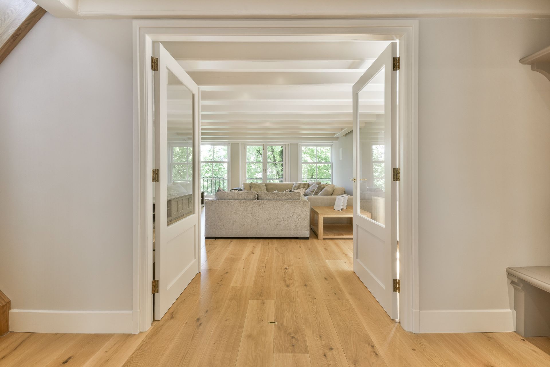 Open white glass doors reveal a living room with light wooden floors, a neutral sofa, and windows.