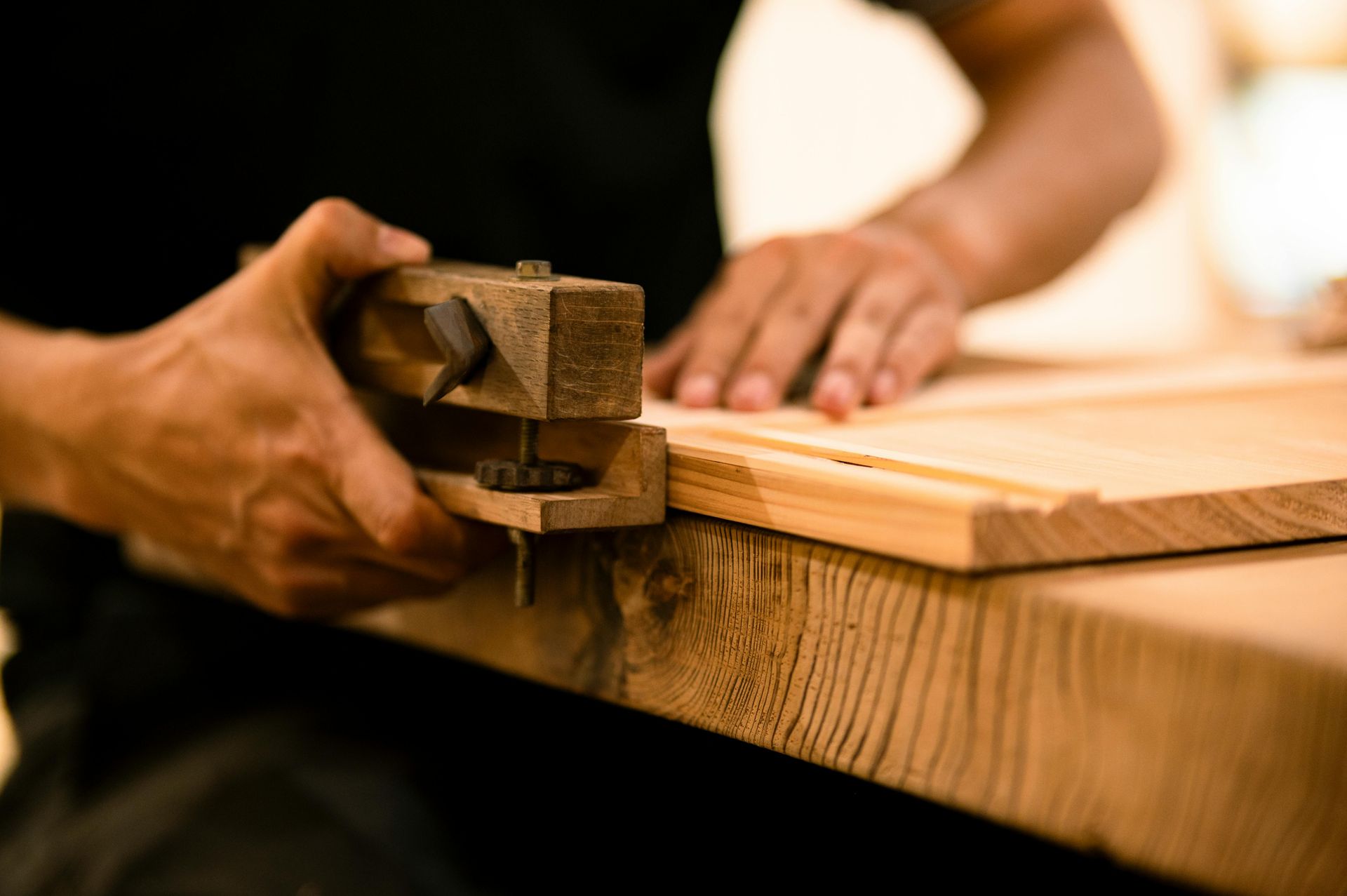 A close-up of hands using a wooden marking gauge to measure a piece of timber on a workbench.