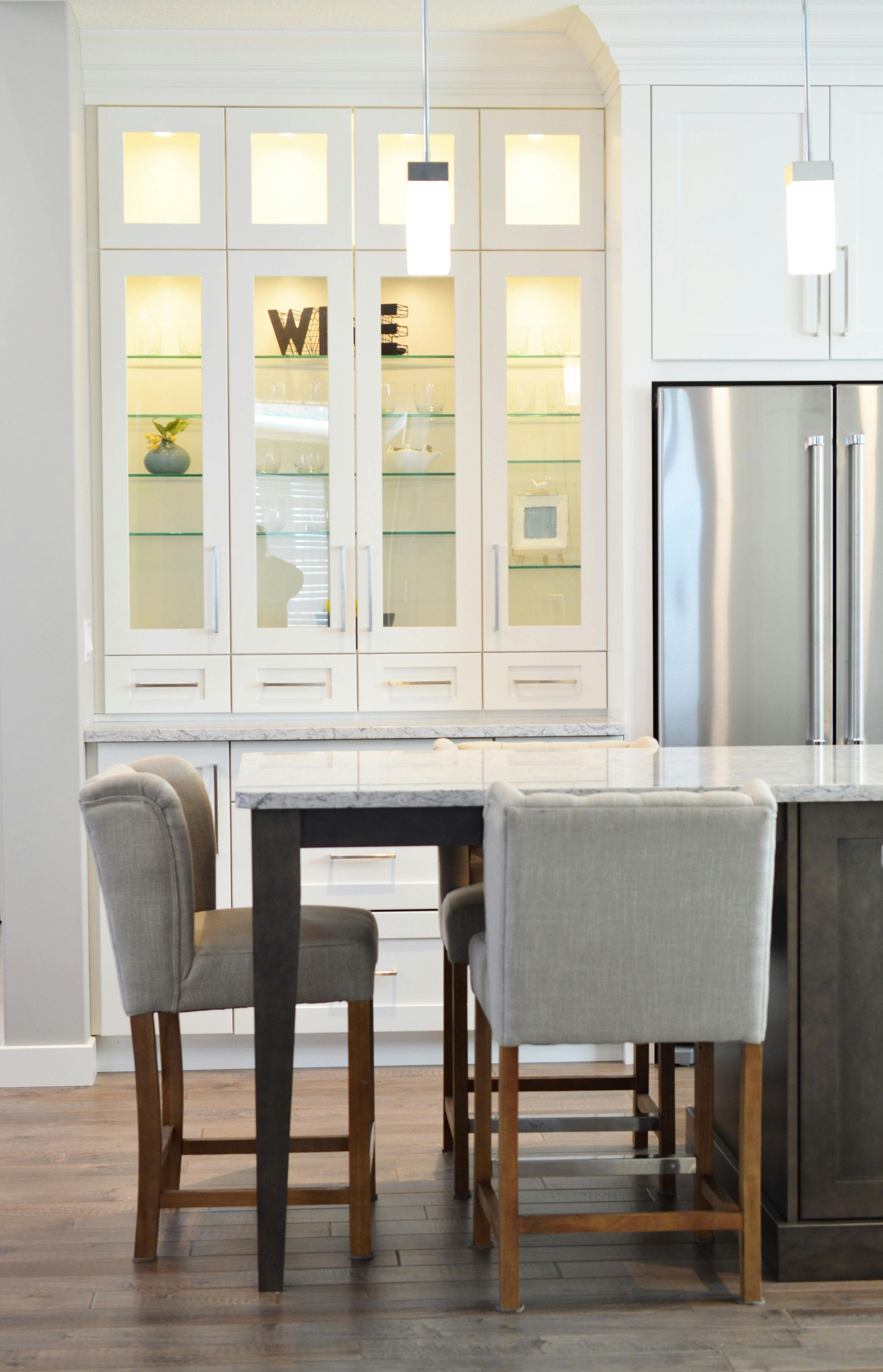 Kitchen with white cabinets, glass-front display, and stainless steel fridge. Bar stools sit at an island and a table.