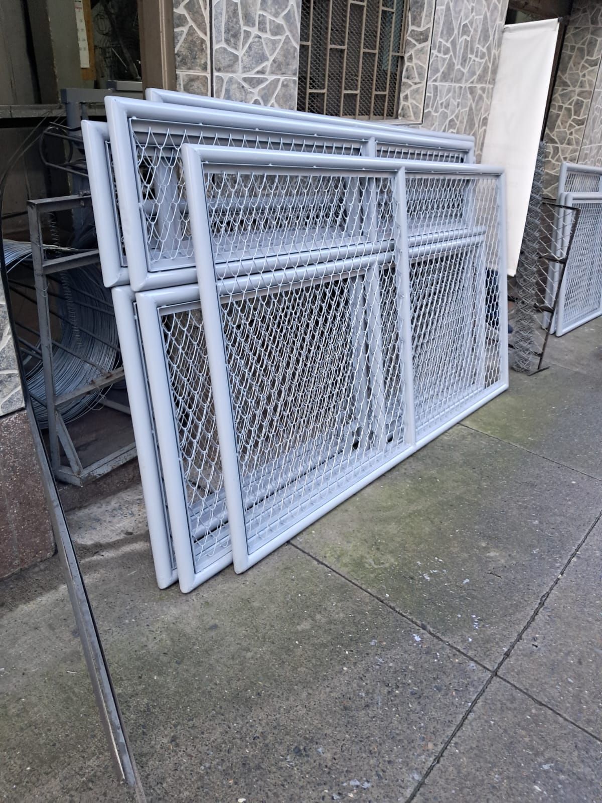 Puertas apiladas de malla metálica blanca apoyadas contra una pared al aire libre, en un camino de hormigón.
