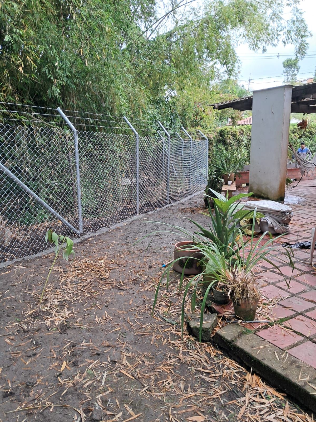 Una valla de tela metálica bordea un sendero de grava frondoso con macetas y un patio embaldosado.