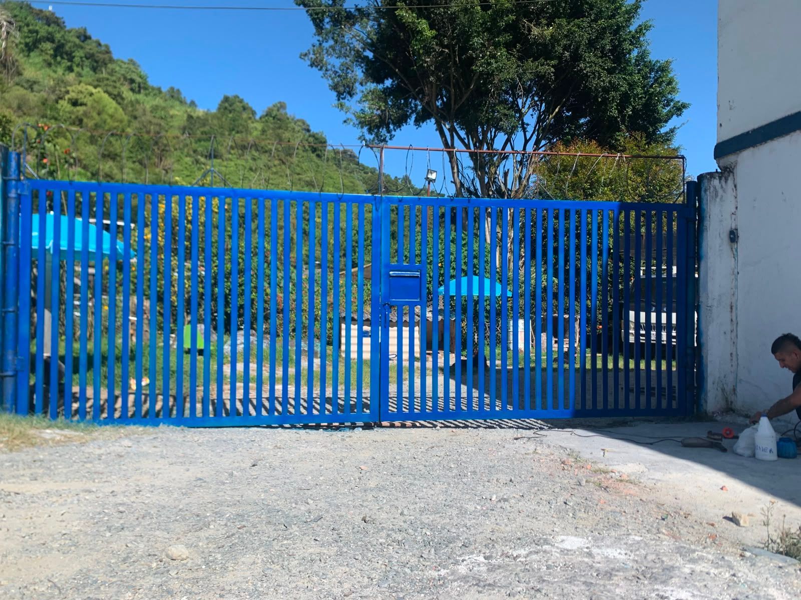 Una puerta metálica azul cruza un camino de entrada con colinas verdes y árboles al fondo