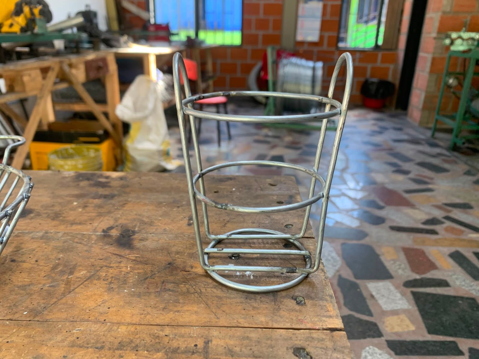 Cesta de alambre metálico sobre una mesa de madera en un taller con suelo de baldosas y sillas al fondo.