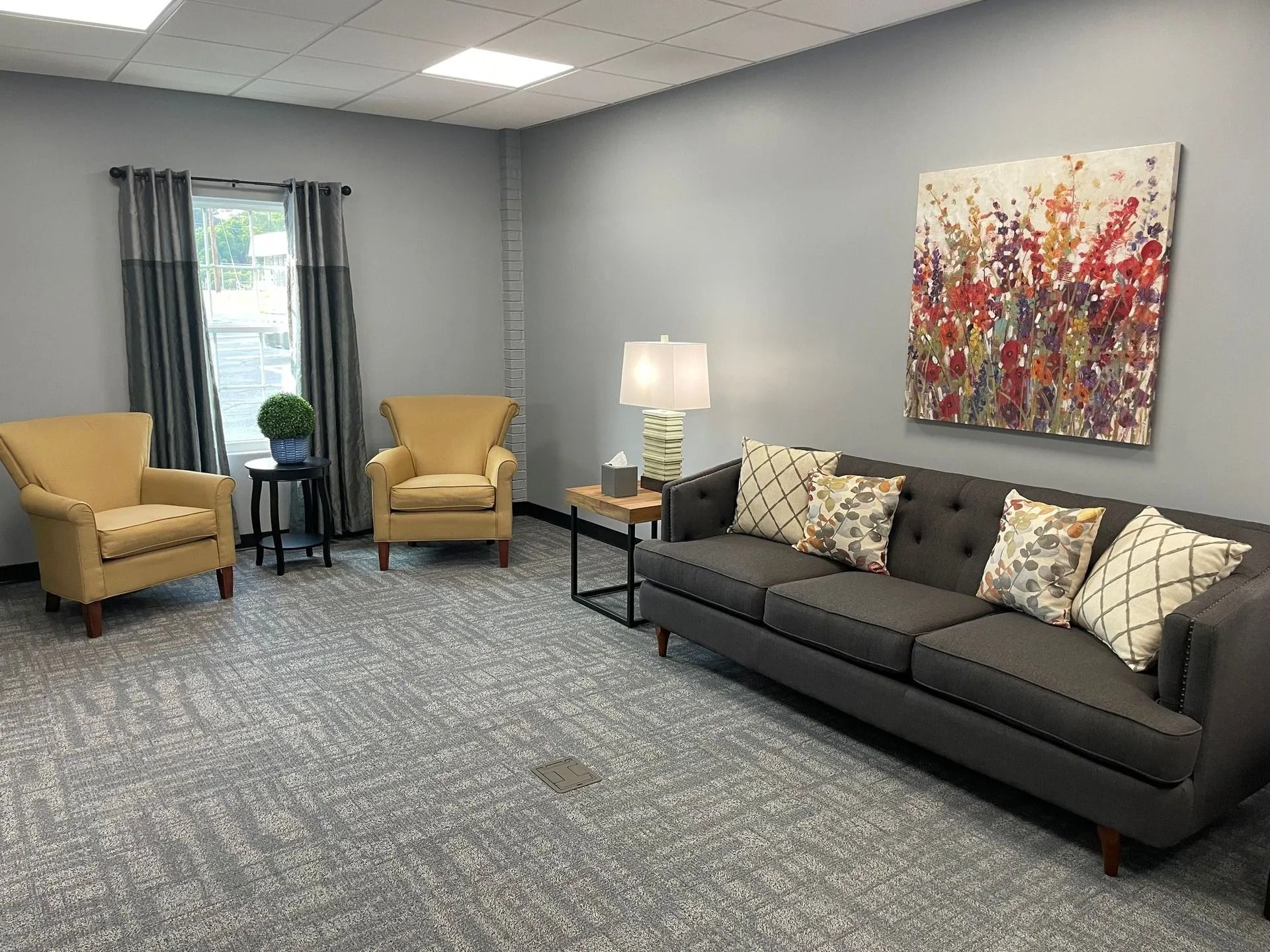 Waiting room with gray sofa, yellow armchairs, artwork, and lamp.