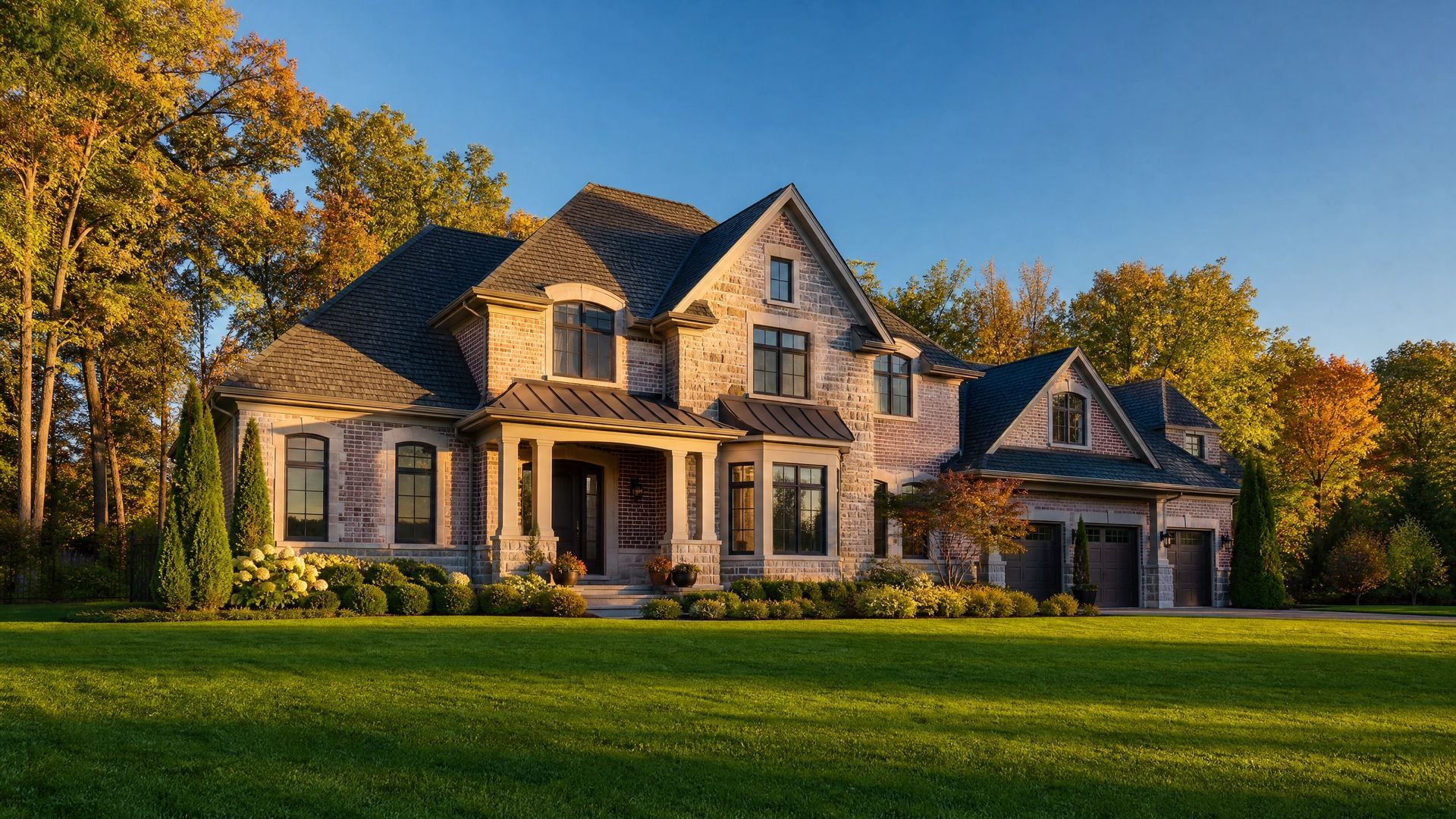 Large stone house with a front porch and garage, set on a green lawn with autumn trees at sunset