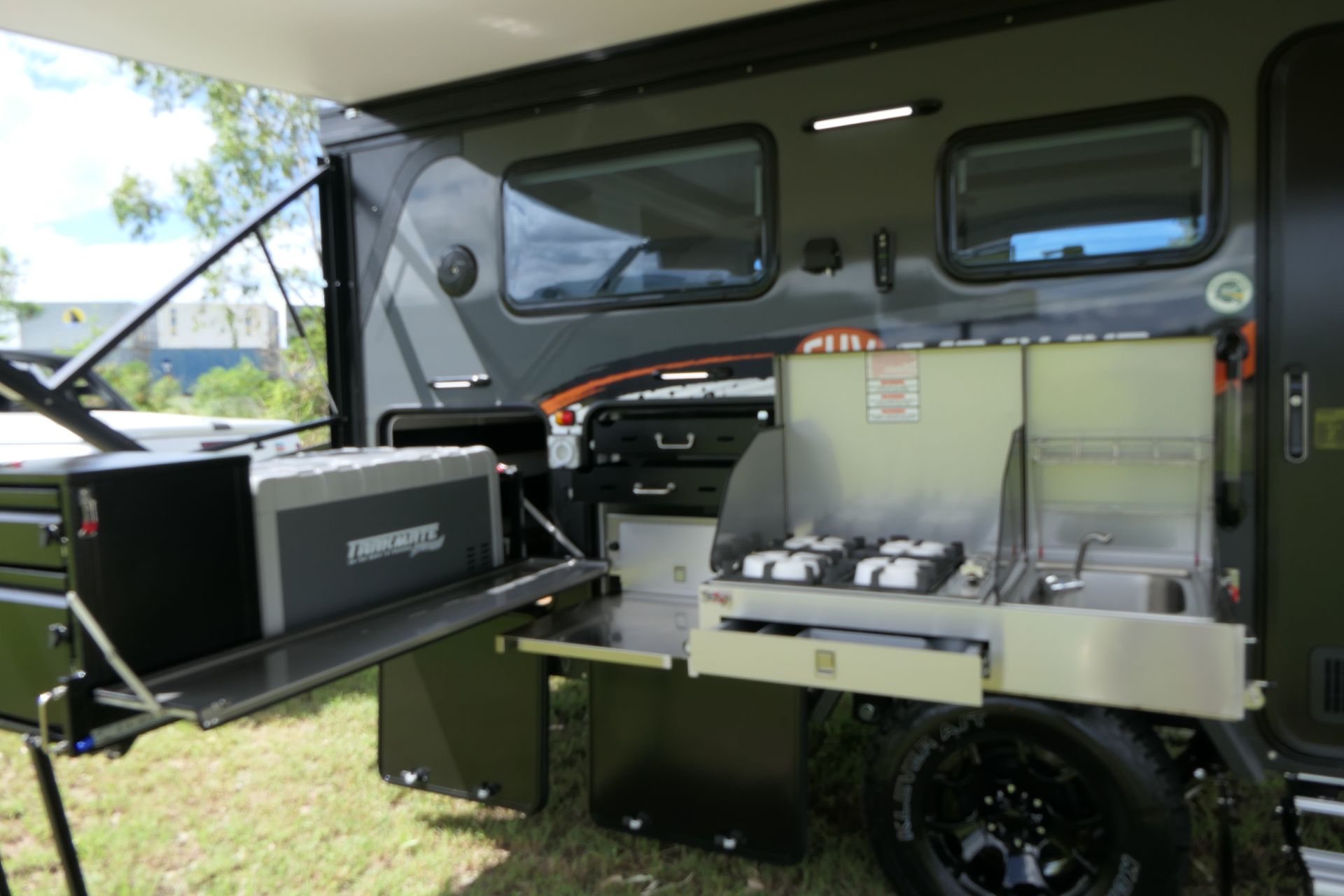 Camper trailer with outdoor kitchen, fridge, and awning. Dark green and black color scheme.