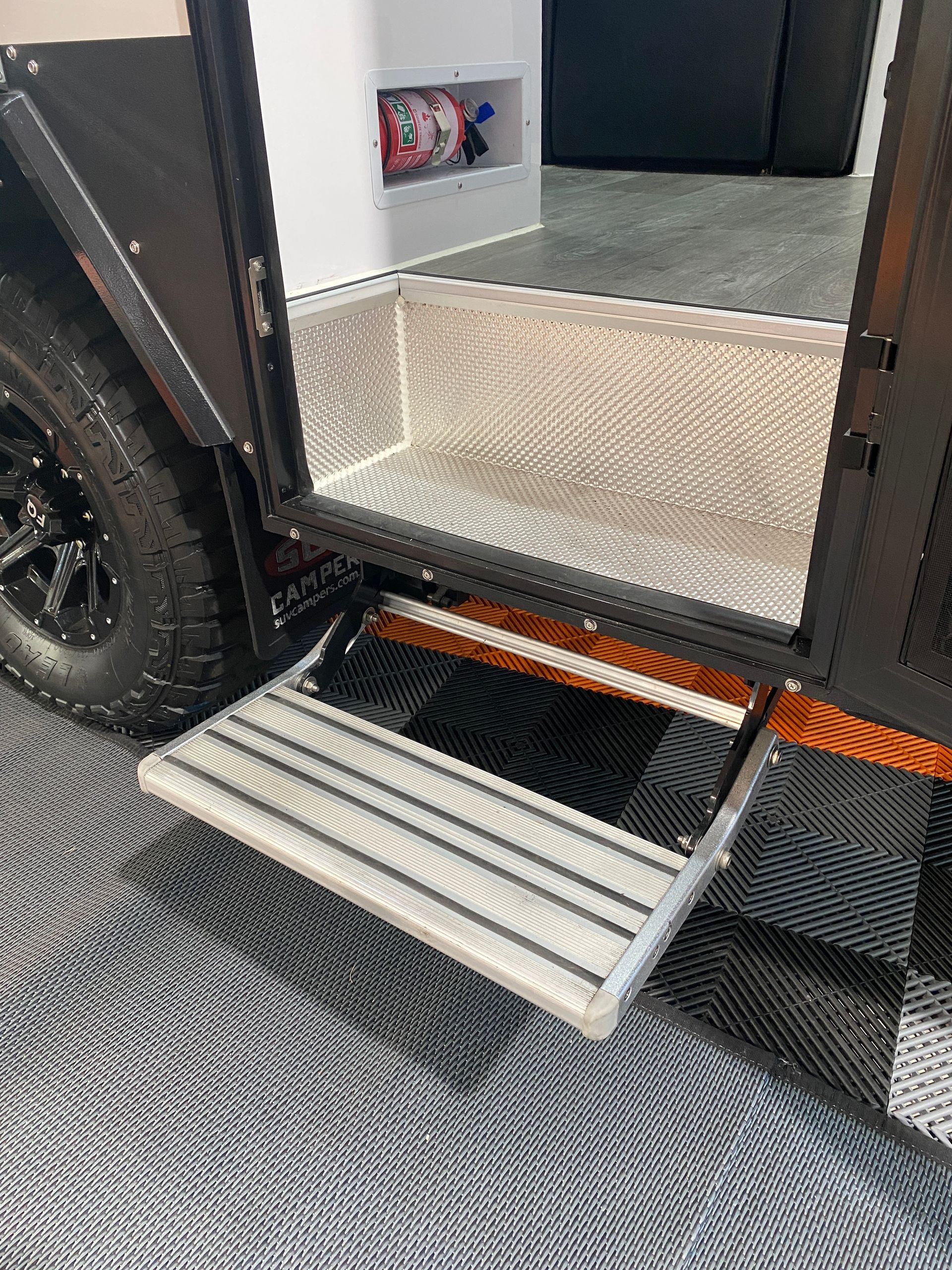 Open RV doorway with extendable step. Aluminum steps and interior, dark frame, tire visible.