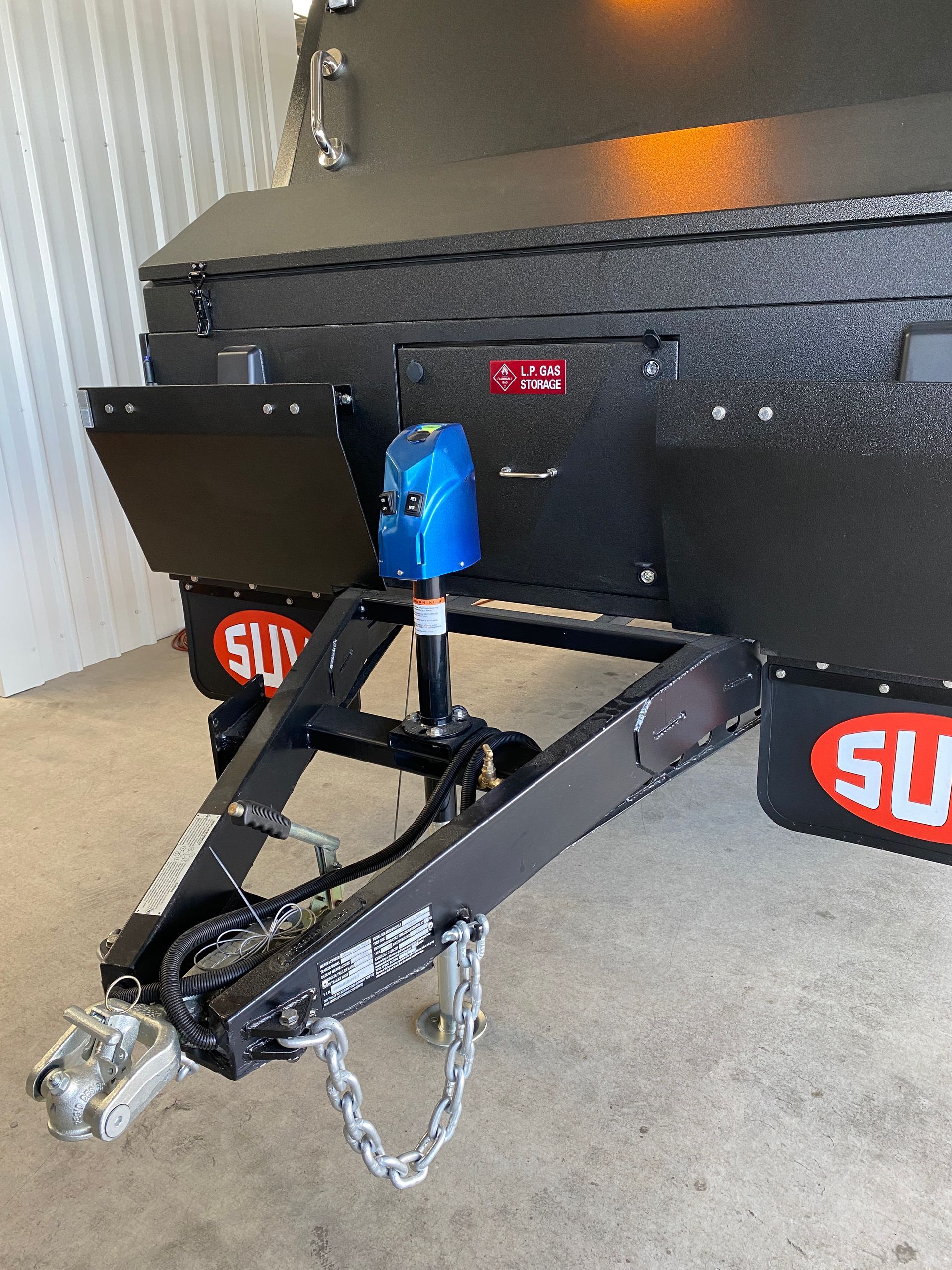 Black trailer with a blue jack, mounted on a hitch. The trailer has black storage compartments.