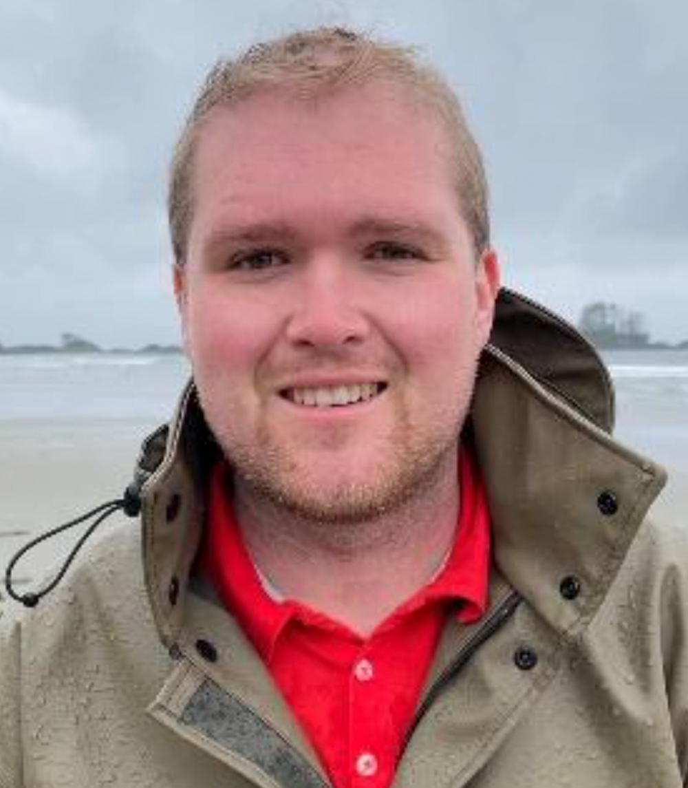 Un homme portant une veste et une chemise rouge sourit sur une plage.