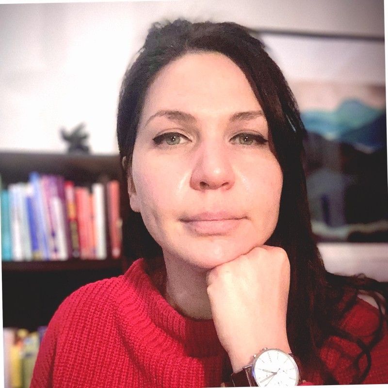 A woman wearing a red sweater and a watch is standing in front of a bookshelf.