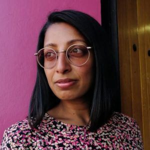 A woman wearing glasses is standing in front of a pink wall.