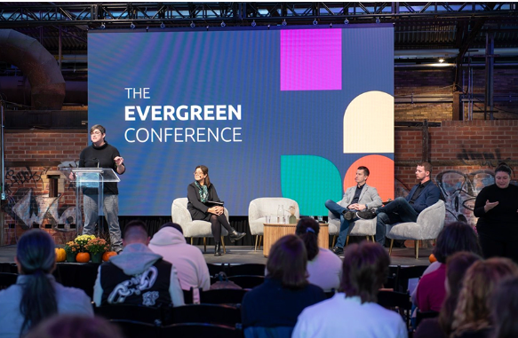A group of people are sitting in front of a large screen that says the evergreen conference.