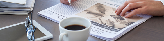 A person is reading a newspaper next to a cup of coffee.