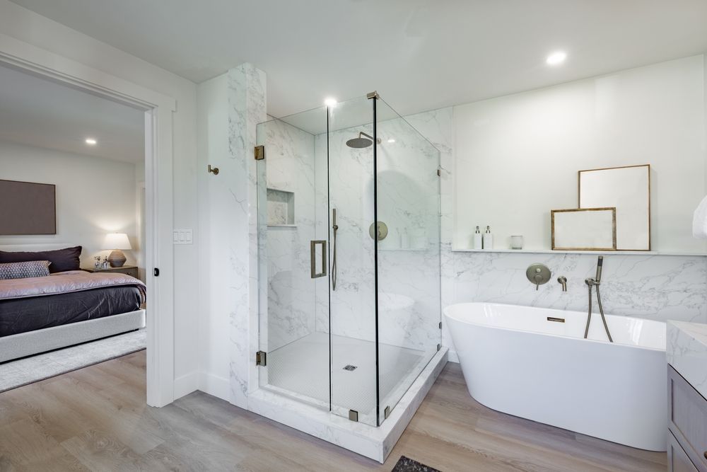 Bright modern bathroom with glass shower, freestanding tub, white marble walls, and bedroom visible nearby