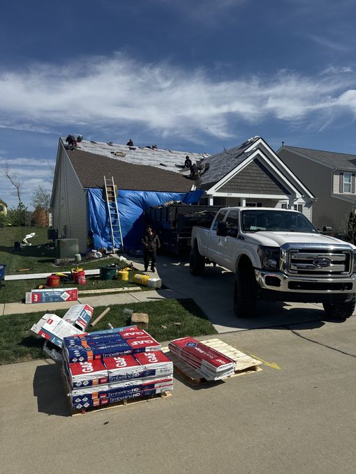 Roofing supplies and pickup truck outside a house under repair, with blue tarp on the roof and materials in the driveway