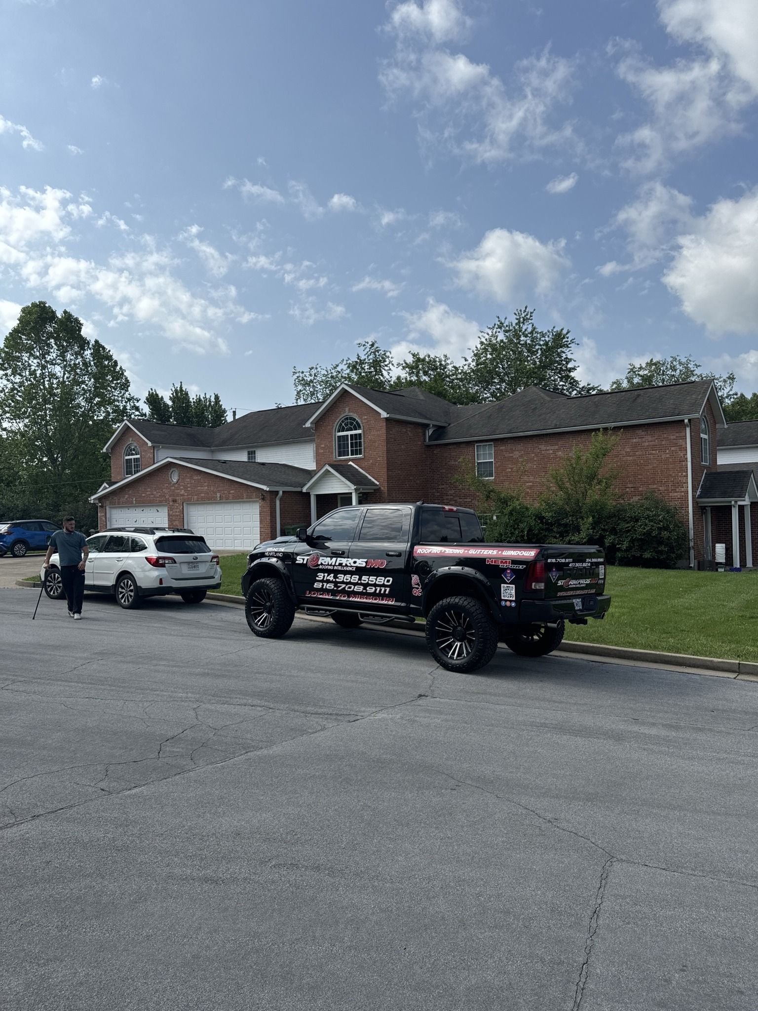 Street view with a lifted black truck, white SUV, and brick houses under a cloudy sky