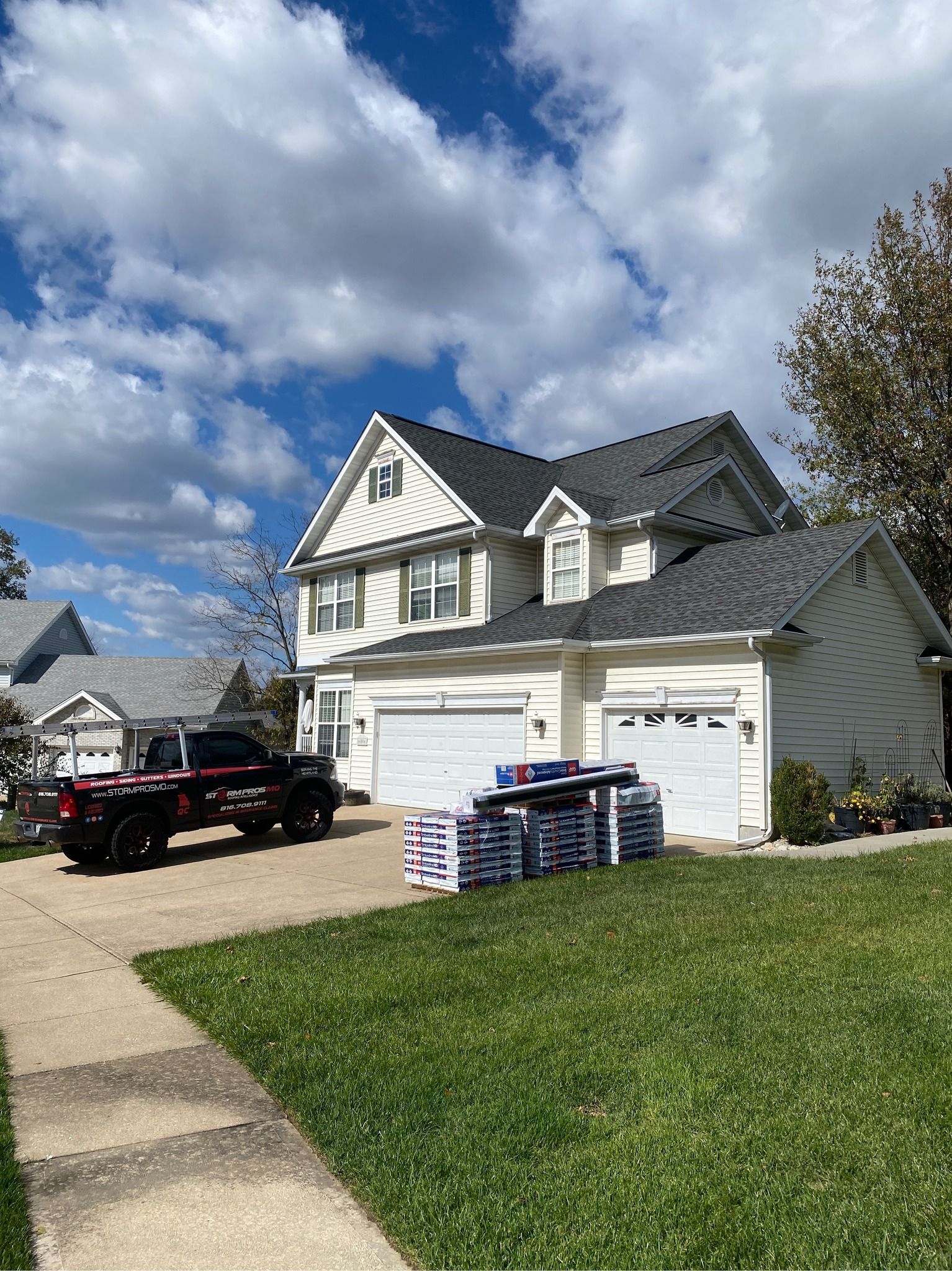 Suburban house with white garage, black truck in driveway, and stacked boxes on the lawn under a cloudy sky