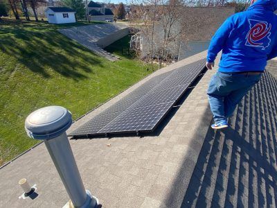 Person on a shingled roof beside a large solar panel and vent, with a lake and houses below.