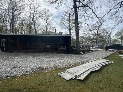 Rustic pavilion beside a gravel lot with bare trees and stacked corrugated metal sheets in the foreground