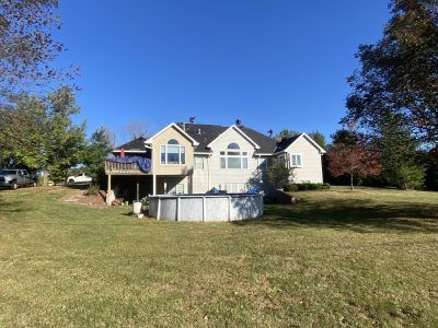 A beige house with a front porch and deck, set on a grassy lawn under a clear blue sky.