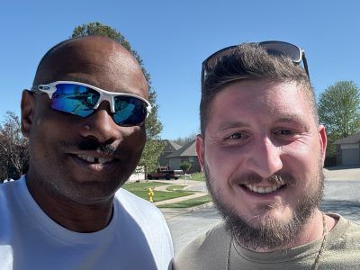Two smiling people outdoors on a sunny street, one wearing sunglasses.