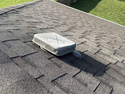 Gray rooftop vent cap on a shingled roof near a grassy edge