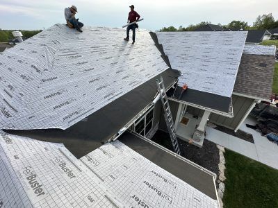 Workers installing roofing underlayment on a house roof with ladders and shingles nearby