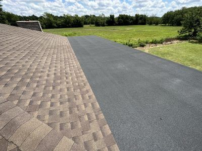 Rooftop with brown shingles and black asphalt roof under a blue sky, bordered by green fields.
