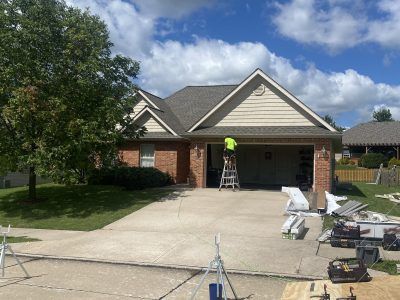 House exterior with open garage, worker on ladder, and tools on driveway under a partly cloudy sky