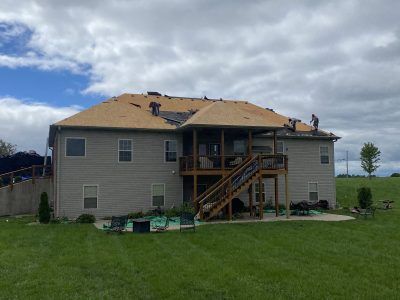 Two-story beige house with wooden deck and stairs in a grassy yard under a cloudy sky