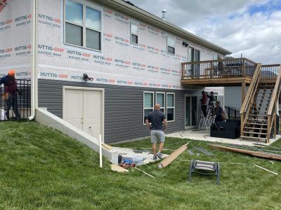 Workers install siding on a house exterior, with scaffolding and a wooden deck staircase nearby.