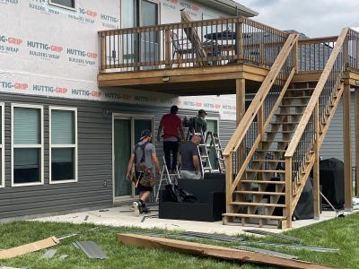 Workers on a house exterior deck and stairs under construction, with siding and scaffolding visible.
