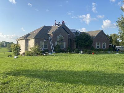 Workers on a ladder repairing a house roof in a grassy yard under a blue sky.