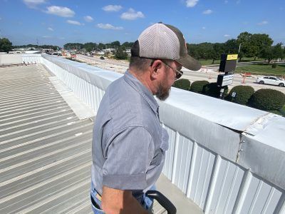 Man in a gray shirt and tan cap leaning over a white railing on a rooftop under a blue sky.