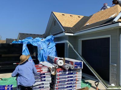 Worker beside stacked roofing materials and blue tarp near a house garage under a clear sky