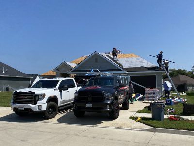Workers reroofing a suburban house with two trucks in the driveway under a clear blue sky
