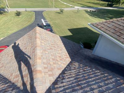 Roof shingles and a dark vehicle parked in a driveway beside a grassy yard on a sunny day