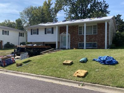 Suburban house with white siding and brick facade; lawn has stacked materials and blue tarp.