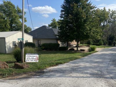 Quiet residential street with a house, trees, and a roadside sign on a sunny day